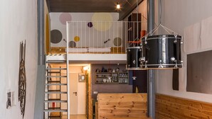Un espacio tipo loft con una mesa de madera, taburetes, una escalera, una zona de cocina y un set de batería.
