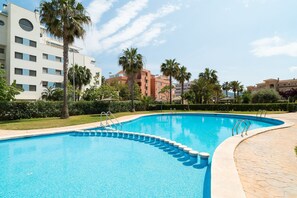 A swimming pool with a curved edge, surrounded by palm trees and a building with multiple floors and balconies. - Fais in D nia With 2 Bedrooms and 2 Bathrooms (Denia)