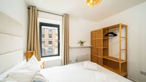 A hotel room with a bed, a window with curtains, a bedside lamp, a wooden wardrobe, and a small plant on a shelf. - Castle Street Apartment - stylish city centre pad (Dundee)