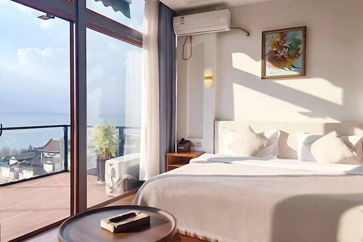 Ein Hotelzimmer mit einem großen Bett, einem Nachttisch, einer Stehlampe, einer Topfpflanze und Blick auf die Stadt durch große Fenster.