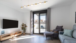 Uma sala de estar moderna com uma televisão de tela plana, um móvel de entretenimento de madeira, um sofá cinza e uma porta de vidro deslizante que dá para uma área externa.