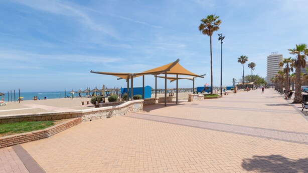 Um calçadão pavimentado com um quiosque coberto, palmeiras e vista para o mar.
