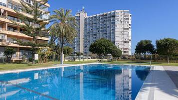 Una piscina con superficie de agua azul cristalina, rodeada por un césped cuidado y árboles. Al fondo, un edificio residencial alto con varios balcones.