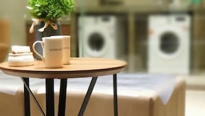 Taula rodona petita amb una tassa de cafè i un recipient petit a sobre, una planta en test i un llit amb estovatge blanc al fons.
