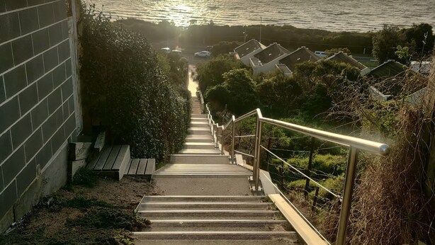 Eine Treppe führt zu einer Küstenlandschaft mit Blick auf das Meer und einen untergehenden Sonnenuntergang.