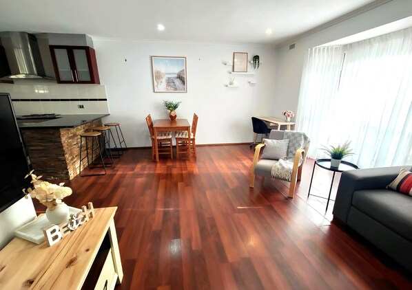 A modern living room with a wooden floor, a dining area with a table and chairs, a sofa, and a kitchen area with a stove and cabinets.