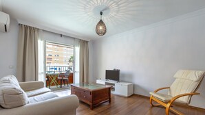 Un soggiorno moderno con un divano, un tavolino in vetro, una poltrona a dondolo in legno, una TV su un mobile bianco e un'ampia finestra con tende.