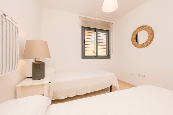 A bedroom with two beds, a bedside table with a lamp, a window, and a wall-mounted mirror.