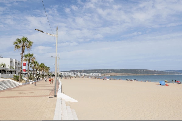 A beach with a paved walkway, palm trees, and beach umbrellas. - La Yerbabuena in Barbate (Barbate)