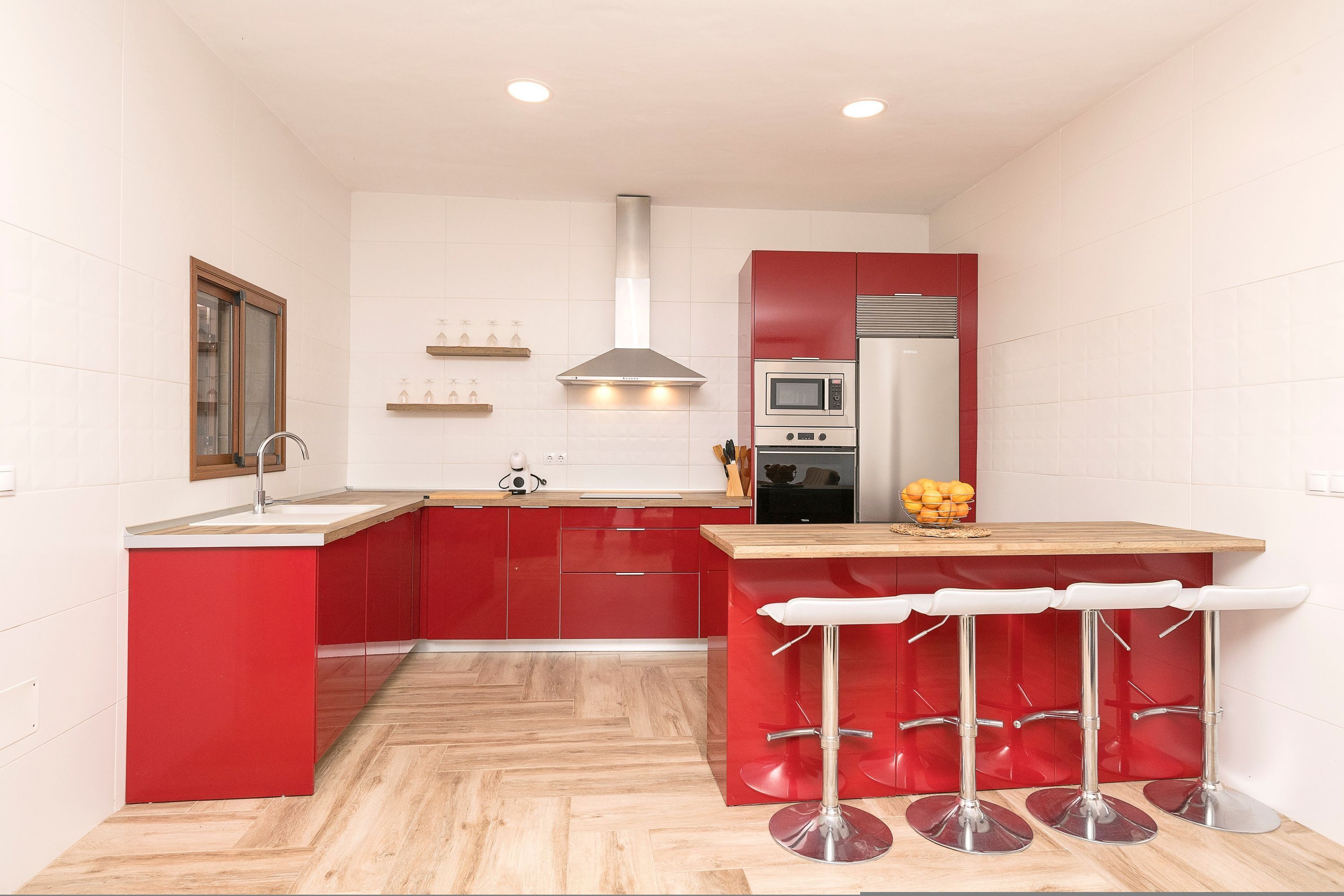 Une cuisine compacte avec des meubles rouges, un plan de travail en bois, une hotte de cuisinière en acier inoxydable, un four à micro-ondes et un réfrigérateur. Il y a trois tabourets de bar blancs devant le comptoir.