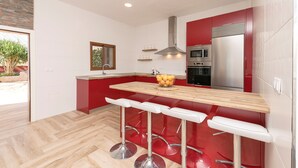 Una cocina moderna con armarios rojos, encimera de madera y electrodomésticos de acero inoxidable. Hay tres taburetes blancos junto a una ventana con vistas a zonas verdes.