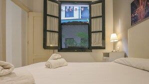 A hotel room with a bed, a nightstand with a lamp, a window with curtains, and a towel on the bed. - Patio De La Cartuja 4 Pax (Seville)
