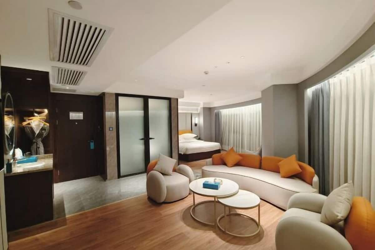 Quarto de hotel moderno com sofá, poltronas, mesa de centro, cama e banheiro com chuveiro.
