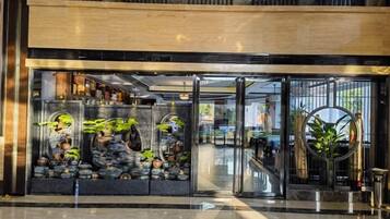 Entrada de hotel con fachada de vidrio, un mostrador de recepción y un arreglo de plantas decorativas.