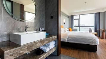 Una habitación de hotel moderna con un amplio tocador de baño, una cama con ropa blanca y un gran ventanal con vistas a la ciudad.