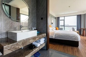 A modern hotel room with a large bathroom vanity, a bed with white bedding, and a large window offering a city view.