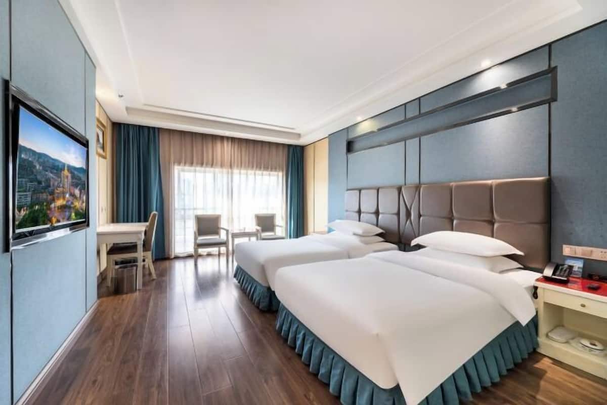 Quarto de hotel com duas camas, uma mesa de escritório, cadeiras, televisão e uma janela grande com cortinas.