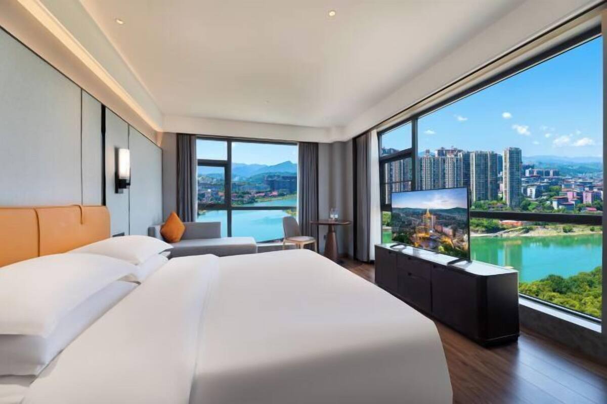 Quarto de hotel moderno com uma cama grande, TV e vista para a cidade através de amplas janelas.