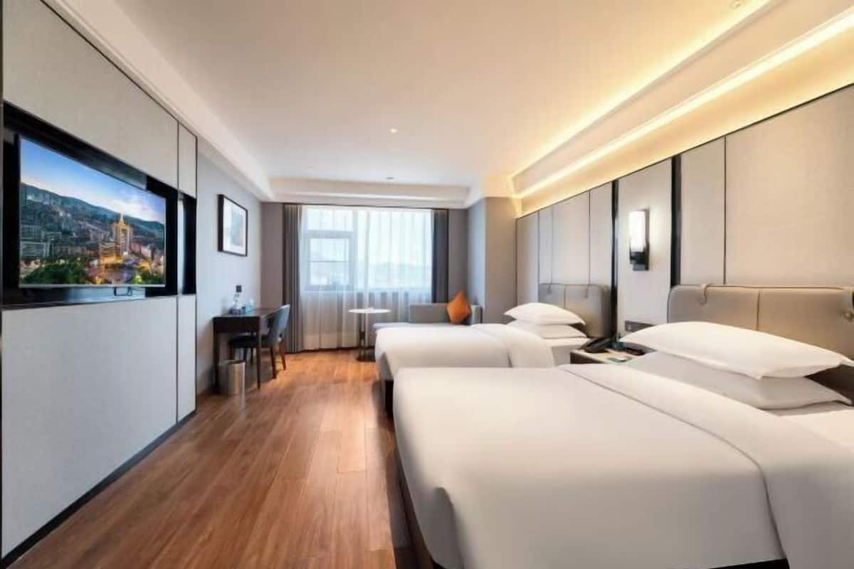 Quarto de hotel com duas camas, uma mesa, uma cadeira, uma televisão e uma janela com cortinas.