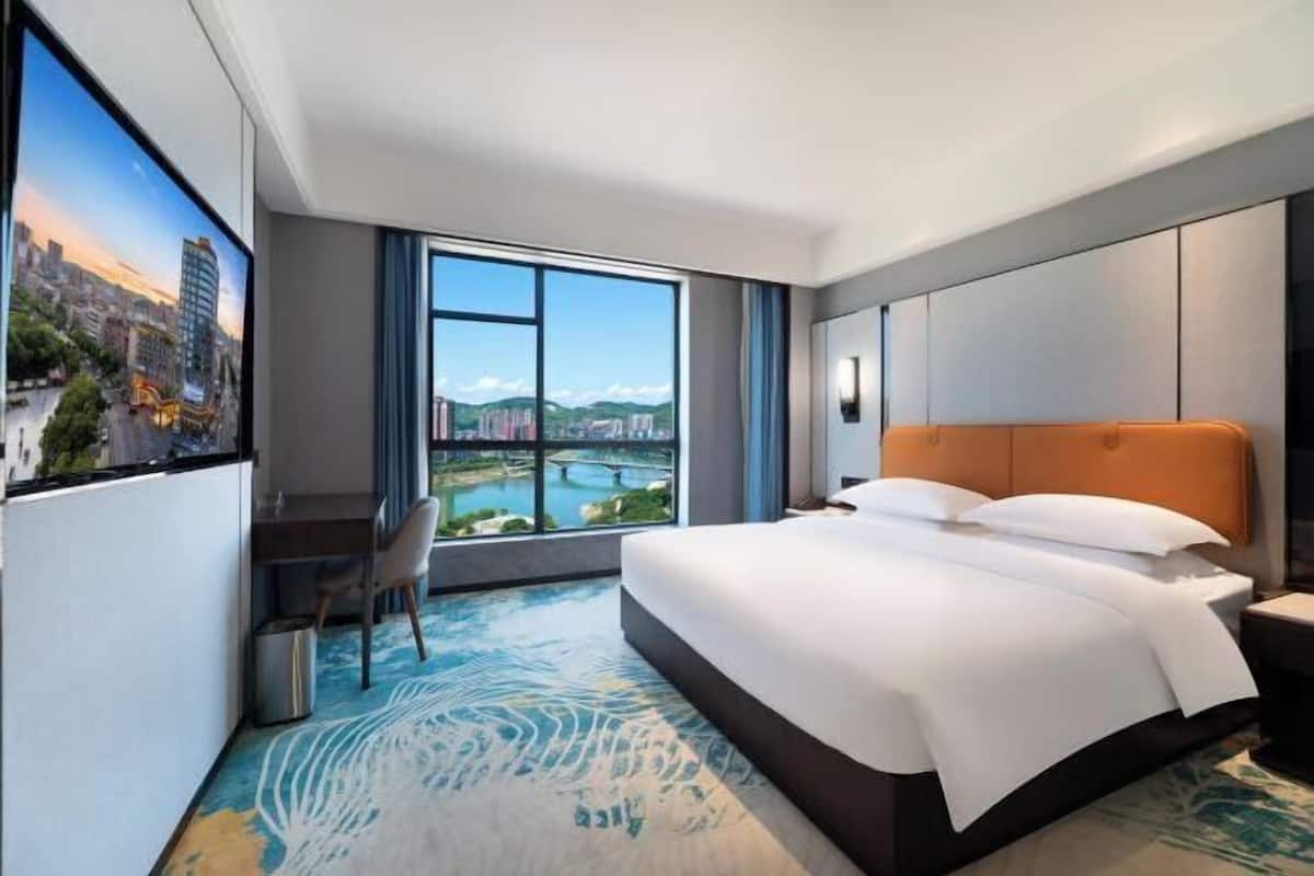 Quarto de hotel com uma cama grande, uma escrivaninha e vista para a cidade através de grandes janelas.