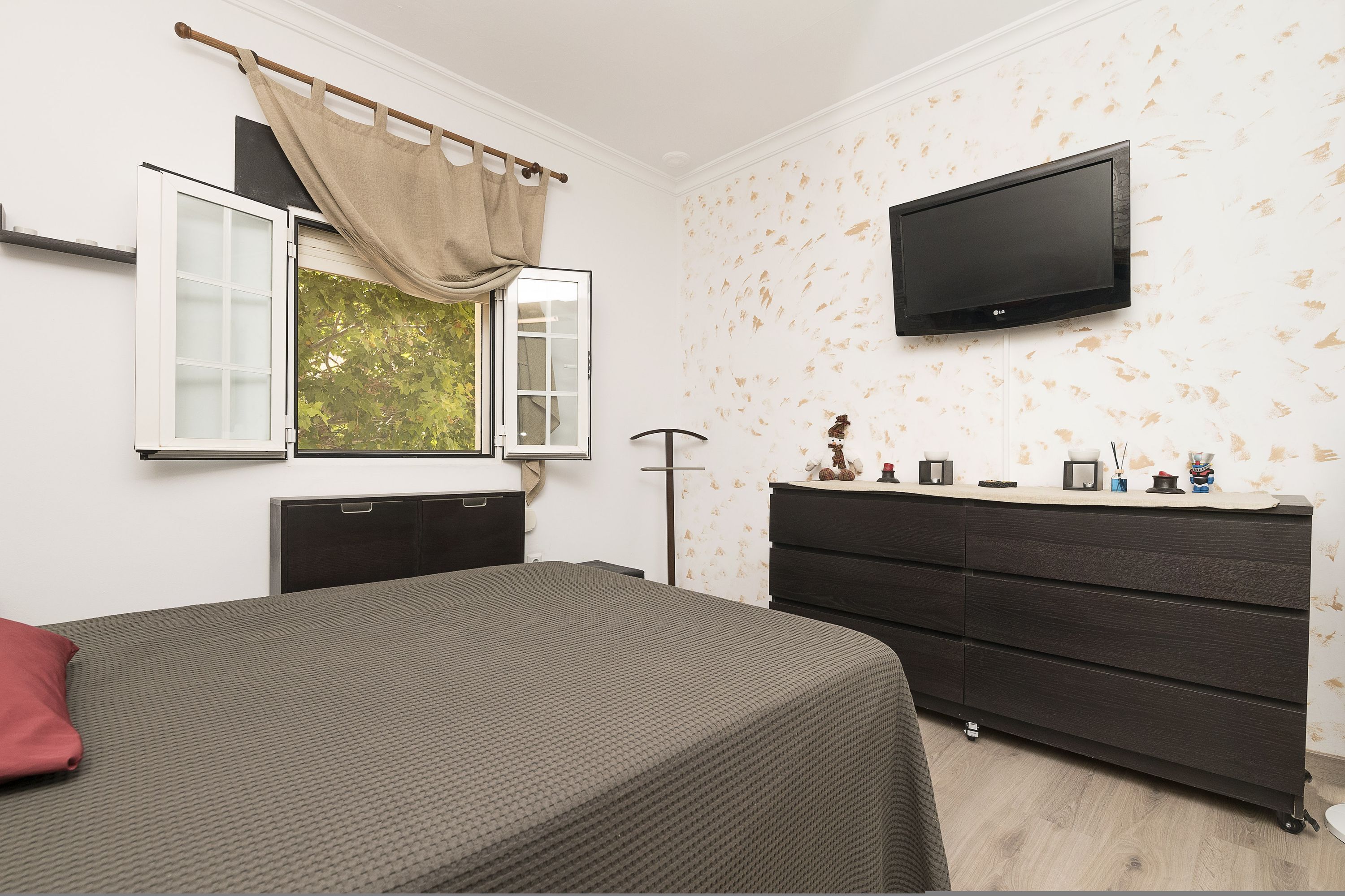 A bedroom with a bed, a dresser, a television mounted on the wall, and a window with curtains.