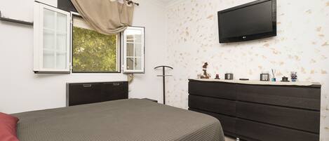 A bedroom with a bed, a dresser, a television mounted on the wall, and a window with curtains.