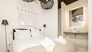 Ein ordentlich bezogenes Bett mit weißen Laken und zwei zusammengerollten Handtüchern, eine Nachttischlampe sowie ein Badezimmer mit Waschbecken und Spiegel.