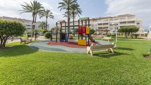 Un parque infantil con una estructura para escalar y toboganes, rodeado de césped y palmeras, con un edificio al fondo.