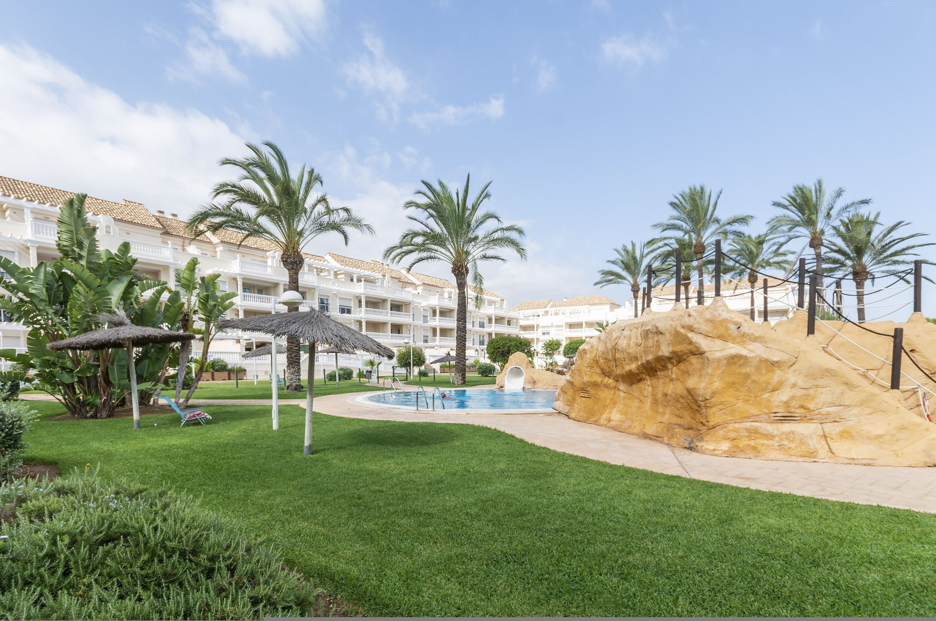 A resort pool area with palm trees, a large rock formation, and a building with balconies in the background.