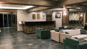 Una hall moderna con una reception, divani verde e beige, un orologio, certificati incorniciati e una grande finestra con vista.