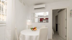 A dining area with a round table, white chairs, and a fruit bowl. There is a window with a curtain, an air conditioning unit, and a mirror above the table. - Retiro Al Amanecer in Chiclana de la Frontera (Chiclana de la Frontera)