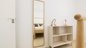 Una habitación con puerta de madera, un espejo grande, un estante de madera con adornos y una cama con cabecera blanca.