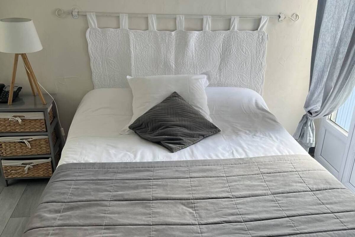 Un lit bien fait, avec une couette grise, des oreillers blancs et un cœur décoratif accroché au mur.