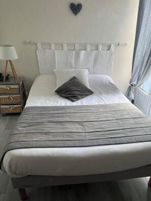 Un lit bien fait, avec une couette grise, des oreillers blancs et un cœur décoratif accroché au mur.