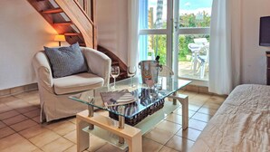 Un salon chaleureux avec une table basse en verre, un canapé et un fauteuil. On y trouve un escalier avec des rampes en bois, une grande fenêtre avec des rideaux et une vue sur l’extérieur.