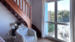 Une pièce avec un escalier en bois, une grande fenêtre avec des rideaux, un fauteuil blanc et une petite table avec une lampe.