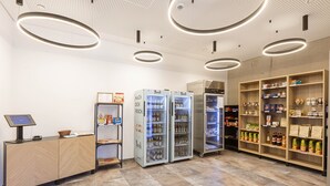 Uma loja de conveniência limpa e bem iluminada, com caixa de autoatendimento, máquinas de bebidas refrigeradas e prateleiras cheias de lanches e bebidas.