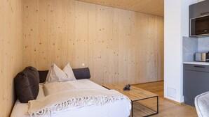 Une chambre avec un plafond en bois, un lit avec une literie blanche, une petite table de chevet avec une plante, et une kitchenette équipée d’un four à micro-ondes et d’une cuisinière.