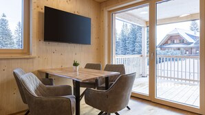 Un intérieur en bois avec un grand téléviseur fixé au mur, une table en bois avec quatre chaises et une porte-fenêtre coulissante donnant sur un balcon d’où l’on peut admirer des arbres couverts de neige et un chalet.