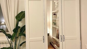 Un armario blanco con dos puertas, una planta a la izquierda y una estantería con objetos decorativos al fondo.