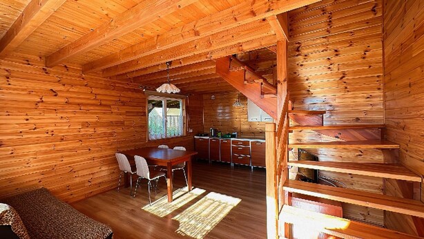 A wooden cabin interior with a dining area, kitchen, and a wooden staircase.