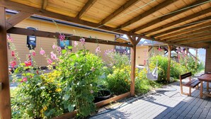 A covered patio with wooden beams, a table and bench, and a garden with various plants and flowers.