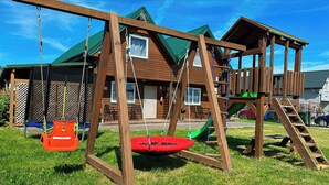 A wooden play structure with swings and a slide, set in a grassy area with a house in the background.