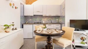 Une cuisine compacte avec des meubles blancs, une petite table à manger avec des chaises, un téléviseur et une arche en briques au-dessus de l’évier.