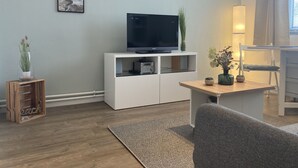 A modern living room with a flat-screen TV on a white cabinet, a wooden side table with a lamp, and a grey sofa. - A little time out for relaxing days... Close to the beach and the center! (Dahme)