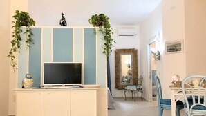 Uma sala de estar com uma TV sobre um móvel branco, uma mesa de jantar com cadeiras e um espelho na parede.
