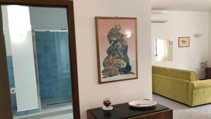 Ett rum med en glasdörr som leder till en duschhörna, ett inramat konstverk på väggen, ett litet bord med en tallrik och en burk, och en gul soffa.