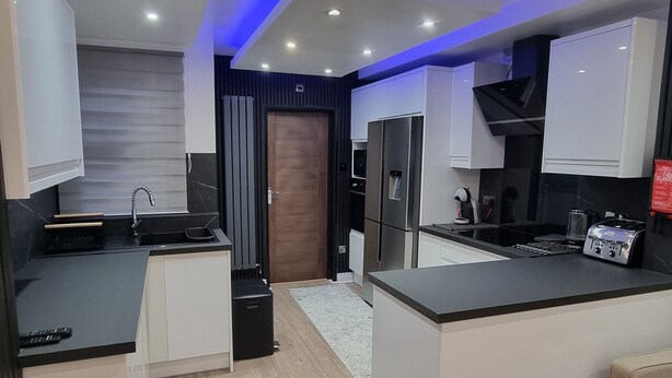 A modern kitchen with white cabinets, a black countertop, and a built-in oven. There is a wooden door and a fire extinguisher on the floor. - Enjoyable 3-bedroom house with WiFi in Watford central / 30min to Central London (Watford)