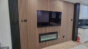 A modern living room with a flat-screen TV mounted on a wooden panel wall, a built-in fireplace, and a kitchen area visible to the right. - Enjoyable 3-bedroom house with WiFi in Watford central / 30min to Central London (Watford)