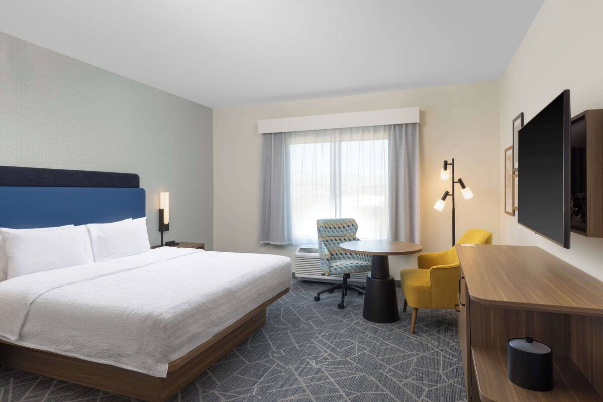 Habitación de hotel con una cama grande, un escritorio, una silla, una mesita y un televisor montado en la pared.